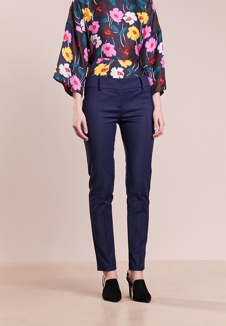 Femme portant un pantalon slim bleu marine, des chaussures à talons noirs et un chemisier sombre à fleurs roses, jaunes et blanches.