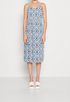 Vrouw staand, gekleed in een mouwloze midi-jurk met blauw-witte geometrische patronen en witte platform slippers.