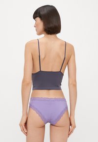 Šedý přiléhavý crop top s tenkými ramínky doplněný levandulovými bikinovými kalhotkami s krajkovým lemem, které vystavují hladkou látku a jemné detaily.