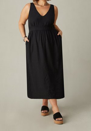Schwarzes, ärmelloses Maxikleid mit V-Ausschnitt, gerafftem Taillenbereich und Seitentaschen. Kombiniert mit schwarzen Wildleder-Plateausandalen. Glatte Textur.