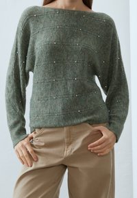 Grön stickad tröja med bred halsringning, prydd med texturerade ränder och spridda silverdetaljer, matchad med beiga byxor.