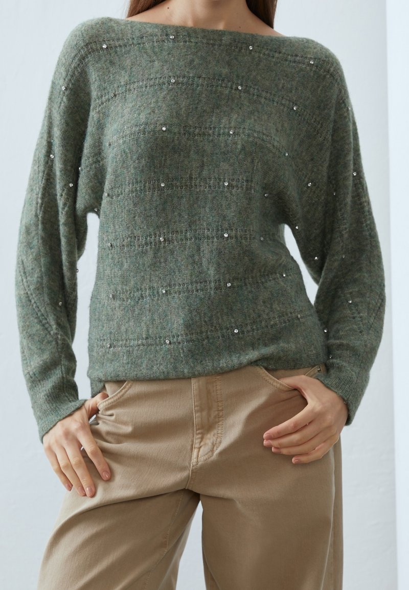 Grön stickad tröja med bred halsringning, prydd med texturerade ränder och spridda silverdetaljer, matchad med beiga byxor.