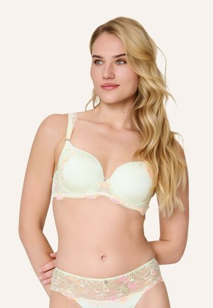 Femme blonde portant un soutien-gorge en dentelle florale pastel et une lingerie assortie en dentelle, regardant légèrement vers la droite sur un fond clair uni.