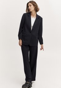 Navy blue blazer with notched lapels and two front pockets, paired with matching trousers. Worn over a white top, featuring a lace neckline.