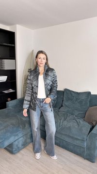 Quilted mørkegrønn jakke med stående krage, kombinert med lyse jeans og joggesko. Scenen er satt i en stue med en sofa.