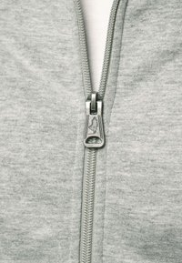 Közeli kép egy ezüst cipzárról világosszürke anyagon, a cipzár húzóján egy kis dombornyomott figura látható, amelyen egy kalapot viselő ember látható.