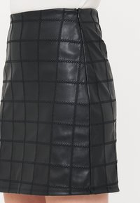 Jupe noire en cuir synthétique avec un motif en damier et des coutures en relief. Fermeture éclair sur le côté et une silhouette droite qui se rétrécit au niveau de l'ourlet.