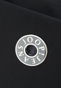 Sort stof med et rundt metalaccent med indgraveret tekst, der læser "JOOP! JEANS." Teksturen er glat med en blank finish.