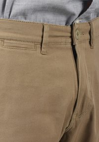 Pantalones de algodón en color beige con una textura suave, bolsillos frontales planos, cierre de cremallera y un solo botón. La costura es visible a lo largo de las costuras.