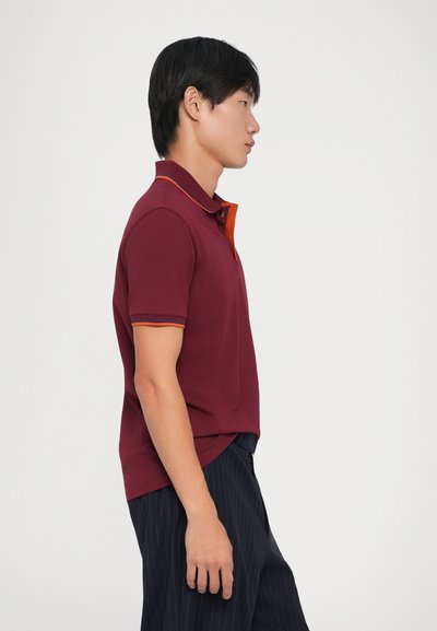 Μπορντό πόλο μπλουζάκι με κοντά μανίκια, πορτοκαλί γιακά και λεπτομέρειες, υφασμάτινη υφή, συνδυασμένο με ναυτικό παντελόνι με ρίγες.