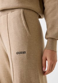 Beżowe spodnie dresowe wykonane z miękkiego materiału, z regulowanym sznurkiem w pasie, bocznymi kieszeniami oraz czarnym haftem "GUESS" na lewym udzie.