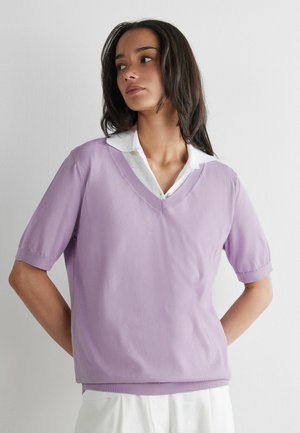 Jeune femme aux cheveux foncés portant un pull violet clair à manches courtes et col en V par-dessus une chemise blanche à col, debout devant un mur uni.