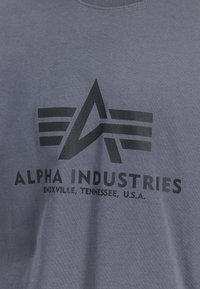 Graues Baumwoll-T-Shirt mit einem schwarzen grafischen Logo, das ein abstraktes Design und den Text enthält: "ALPHA INDUSTRIES, KNOXVILLE, TENNESSEE, U.S.A."