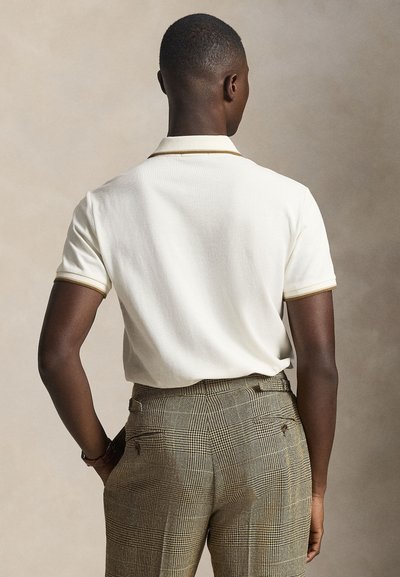 Vyras, atsukti nugara, dėvi trumpomis rankovėmis baltą polo marškinėlį su smėlio spalvos apvadais ir rudas languotas aukštu liemeniu kelnes.