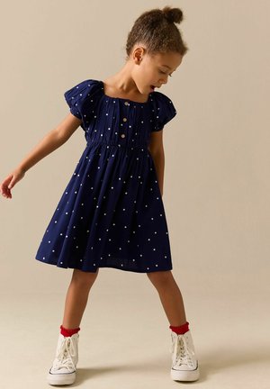 Junges Mädchen in einem marineblauen gepunkteten Kleid, roten Socken und weißen Turnschuhen steht da und blickt nach unten, die Arme leicht ausgestreckt.