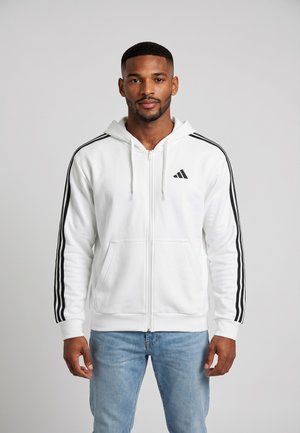 Hombre con sudadera blanca con cremallera y rayas negras en las mangas y vaqueros azul claro, de pie frente a un fondo liso.
