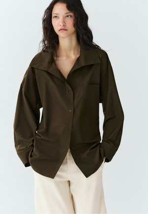 Femme aux longs cheveux foncés portant une chemise ample marron foncé boutonnée et un pantalon beige clair sur un fond clair uni.