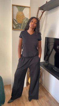Mulher com cabelo encaracolado vestindo uma camisa preta de decote em V e calças largas, está em um ambiente interno perto de uma televisão e uma obra de arte de abacaxi.