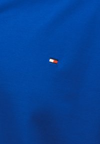 Tissu texturé bleu avec petit logo rectangulaire en rouge, blanc et bleu marine centré sur la surface.