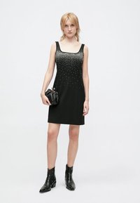 Robe noire sans manches avec décolleté carré et ornements en perles à la taille, associée à des bottines noires et un petit sac à main.