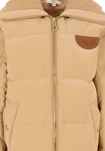 Giacca puffer beige con cerniera dorata e bottoni a pressione. Presenta un colletto a coste e una patch in pelle marrone con logo sul lato sinistro. Texture morbida e trapuntata.