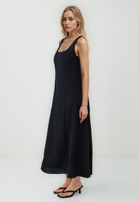 Czarna sukienka bez rękawów z kwadratowym dekoltem i płynącą sylwetką, wykonana z lekkiego materiału, o prostych liniach i długości do połowy łydki.