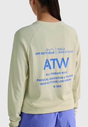 Kvinde iført lysegrøn langærmet nettop med blå "ATW All Terrain Wear" og tilhørende tekst på bagsiden, kombineret med blå bukser.