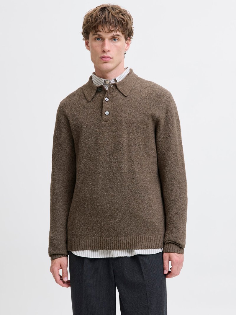 Ung mann kledd i en brun teksturert langermet genser over en stripete skjorte med knapper og mørkegrå bukser, stående mot en hvit bakgrunn.