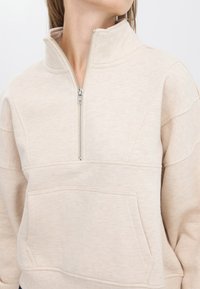 Pull-over beige à zip quart, fabriqué en tissu doux, avec une grande poche avant et une texture subtile. La fermeture éclair est de couleur argentée.