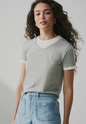 Young woman with curly hair wears layered white and gray short-sleeve shirts and light blue high-waisted jeans with front pockets.