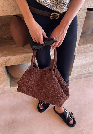 Kobieta trzyma plecioną brązową torebkę i czarne okulary przeciwsłoneczne, ma na sobie czarne spodnie, pasek, wzorzysty top i czarne sandały na klamrę, stoi w pomieszczeniu.