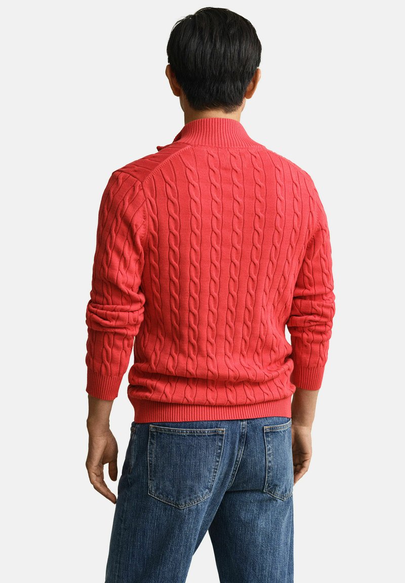 Uomo visto di spalle che indossa un maglione rosso a trecce e jeans blu, su uno sfondo chiaro e uniforme.