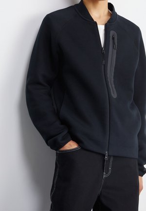 Mand iført sort lynlåsjakke med brystlomme med lynlås og sorte bukser med hvide syninger, hånd i lommen, foran ensfarvet baggrund.