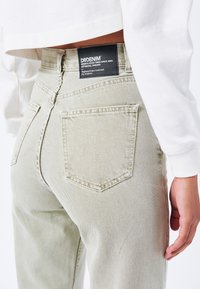 Jeans in denim verde chiaro presentano un design aderente, una texture sottile, tasche posteriori e un'etichetta nera con il marchio in vita.
