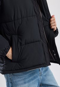 Black padded jacket with quilted design, zip closure, and side pocket. The interior features a contrasting lighter color.