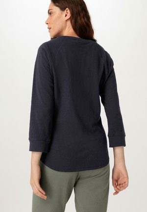 Marineblaues Langarmhemd mit lockerer Passform, Rundhalsausschnitt und geknöpften Ärmeln, kombiniert mit olivgrünen Hosen, die eine weiche Textur aufweisen.