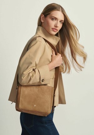 Kobieta o długich blond włosach, ubrana w beżową kurtkę i ciemne dżinsy, trzymająca jasnobrązową zamszową torebkę na ramię z brązowym paskiem.