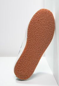 Vit sneaker med en texturerad gummisula. Sulan har ett distinkt mönster, medan sidan har en subtil dekorativ rand.