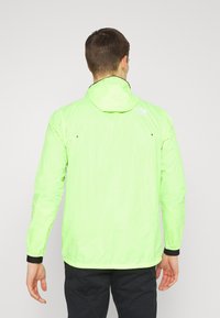 Chaqueta ligera de color verde neón con capucha, mangas largas, puños elásticos y un dobladillo negro. Logo impreso en la parte superior de la espalda. Textura suave.