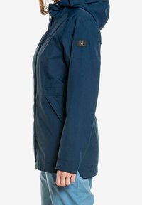 Mörkblå vattentät jacka med huva, utrustad med dragkedja, sidofickor och en logotyp-patch på vänster ärm. Slät textur.