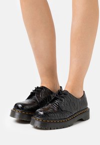 Černé šněrovací boty s texturovaným, vlnitým designem. Silná gumová podrážka s oranžovým akcentem kolem okraje. Hladký, lesklý povrch.