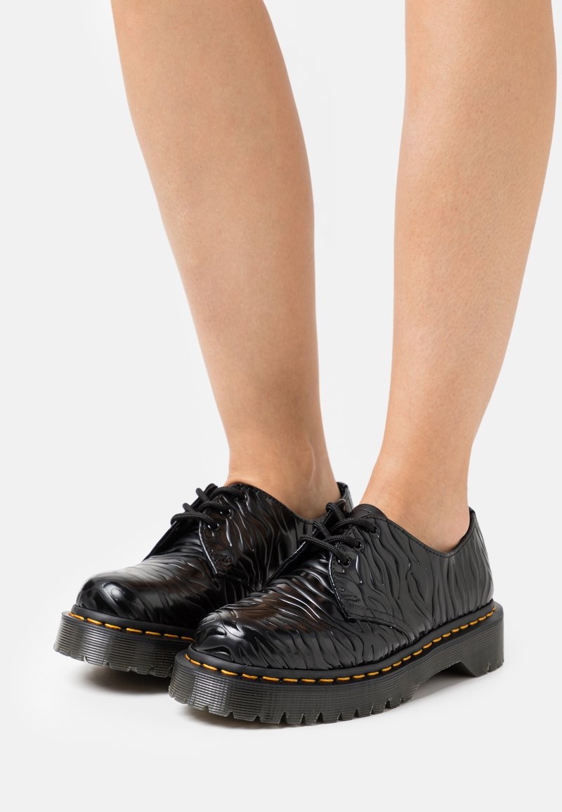 Černé šněrovací boty s texturovaným, vlnitým designem. Silná gumová podrážka s oranžovým akcentem kolem okraje. Hladký, lesklý povrch.