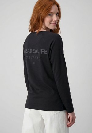 Roodharige vrouw draait zich glimlachend om, draagt een zwart shirt met lange mouwen met "WEAREALIFE" en Japanse tekst op de achterkant, gecombineerd met witte broek.
