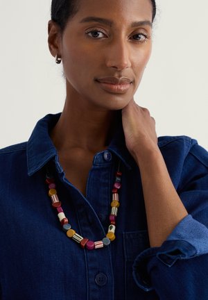 Kvinde med mellemtonet hud iført en mørkeblå denimskjorte og en farverig perlehalskæde, der rører ved sin hals med den ene hånd.