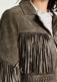 Veste en daim olive avec des franges décoratives et des motifs de feuilles brodés, portée sur une blouse blanche à pois noirs.