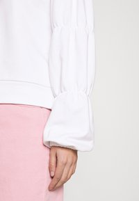 Sweatshirt branco com mangas bufantes, apresentando punhos elásticos. Emparelhado com uma peça de roupa rosa claro, destacando texturas e cores suaves.