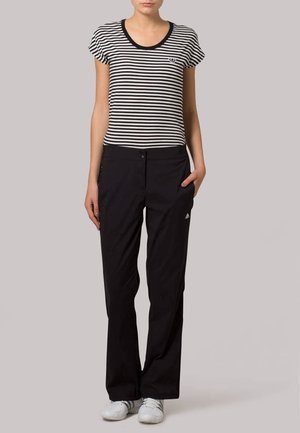 Femme portant un t-shirt à manches courtes rayé noir et blanc, un pantalon noir et des baskets blanches, debout avec une main dans la poche.