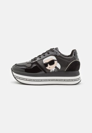 Zapatilla de charol negro con suela gruesa, que presenta una ilustración de un personaje con gafas de sol y acentos de marca a lo largo de la suela.