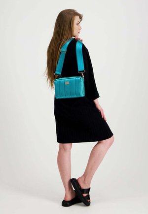 Nainen mustassa mekossa, mustat sandaalit jalassa, kantaa laajakaistaista turkoosia olkalaukkua, seisoo valkoista taustaa vasten.