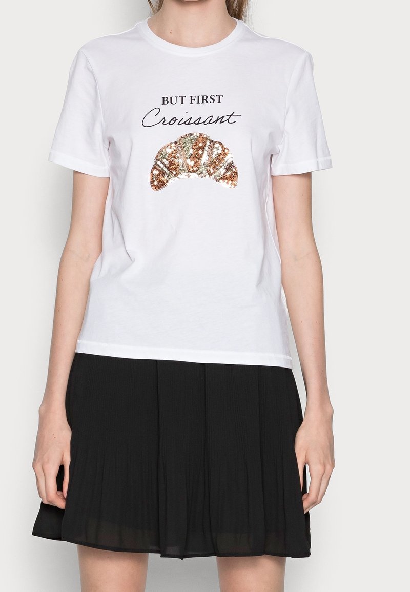 Vit bomulls t-shirt med svart text "MEN FÖRST" ovanför en paljetterad croissantgrafik. Rund halsringning och korta ärmar.